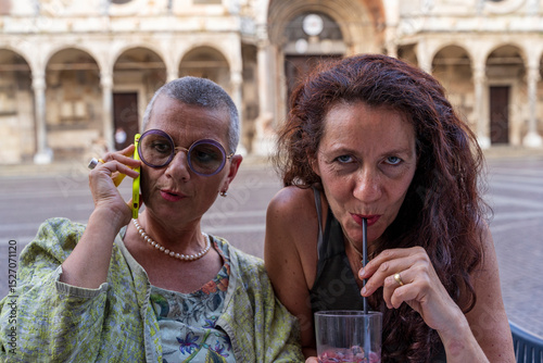 portrait of beautiful middle aged female couple drinking and using phone sitting at outdoor cafe table in historic square
