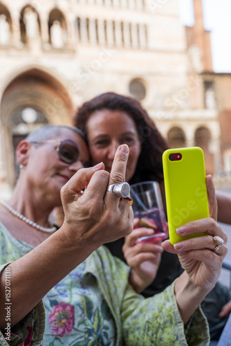 portrait of beautiful middle aged female couple having fun taking a selfie with mobile phone sitting at an outdoor table in a historic square