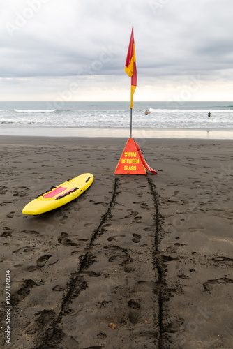 Surf lifesaving flags mark the safe swimming zone on a beach in New Zealand, with a rescue board ready. Swimmers enjoy the water. Karekare, Auckland, New Zealand