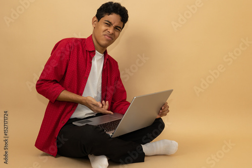 Profile view of young Latino man with a displeased expression wearing red and black casual clothes sitting and pointing with his finger while using his laptop on a light brown background with advertis