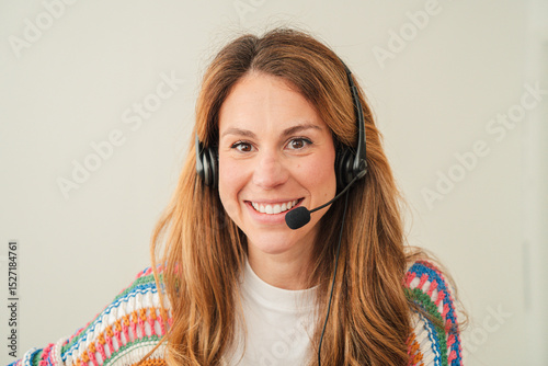 Engaging woman with a friendly smile wearing a headset is focused on her laptop while working in a well-lit home office, showcasing a lively and professional atmosphere for remote communication.