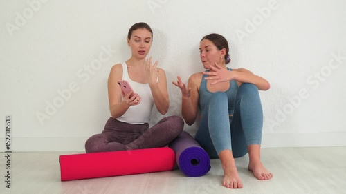 Two smiling women are joyfully engaged in a friendly conversation as they browse their smartphone, sharing fitness tips and wellness ideas while sitting on colorful yoga mats in a relaxed atmosphere