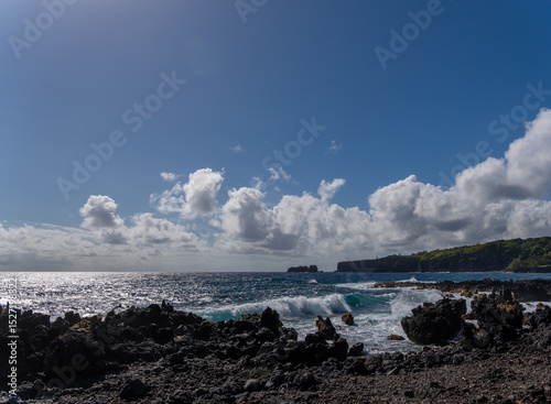 Waves crash on a rocky shore under a blue sky with scattered clouds.