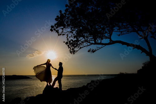 Silueta de pareja bailando al atardecer sobre rocas frente al mar con vestido ondeando al viento
