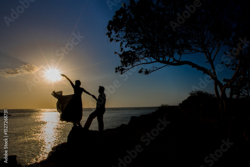 Silueta de pareja bailando al atardecer sobre rocas frente al mar con vestido ondeando al viento
