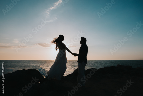 Silueta de pareja bailando al atardecer sobre rocas frente al mar con vestido ondeando al viento

