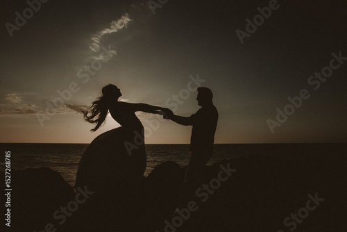 Silueta de pareja bailando al atardecer sobre rocas frente al mar con vestido ondeando al viento
