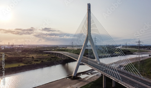Aerial view of the Flintshire Bridge at sunrise, showcasing its elegant cable-stayed design spanning a calm river, with surrounding industrial landscape