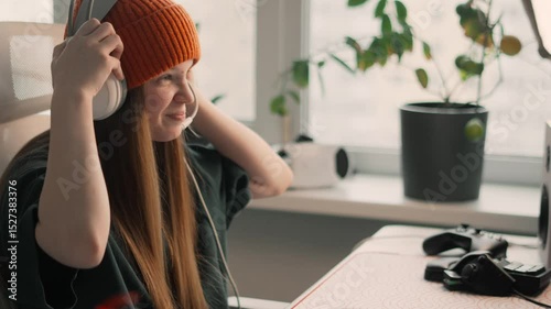 Teenage girl plays online game on desktop while deeply focused on screen. Relaxed posture express modern youth passion for gaming and digital fun. Puts on headphones to communicate with players
