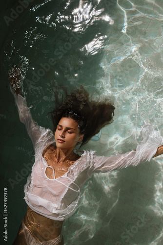 A young woman floats on her back in clear turquoise water, wearing a delicate white shirt. Her face is calm, eyes closed, embodying peace and surrender.