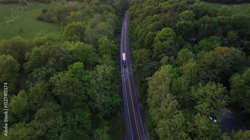 Truck on rural intersection road between green lush trees and farm fields at sunset. Aerial tilt down tracking shot. Idyllic american landscape in United States. Calm Spring evening in USA.