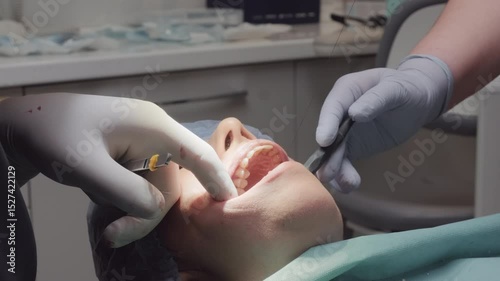 Dental professional works with forceps performing procedure. Assistant manages area with retractor and suction tool ensuring conditions for treatment