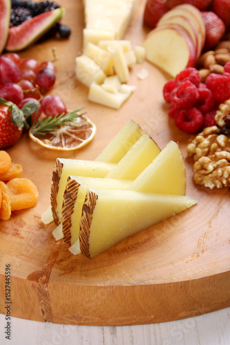 Sliced Manchego cheese on a rustic wooden board accompanied by fresh strawberries, grapes, dried apricots, walnuts, and raspberries