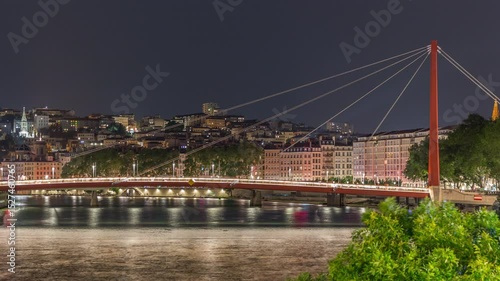 Night timelapse of Lyon's cityscape with the red suspension footbridge over the Saone River, Illuminated waterfront reflected in the water during the evening. France