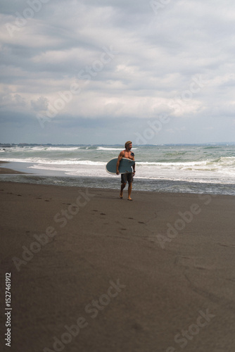 Surfer walking along black sand beach under moody skies