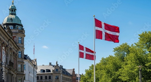 Danish flags flying prominently on flagpoles in a sunny urban street with traditional architecture and green trees.
