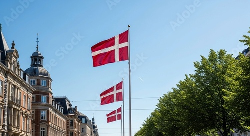 Vibrant Danish flags displayed in an urban setting, featuring historical architecture and bright green foliage.
