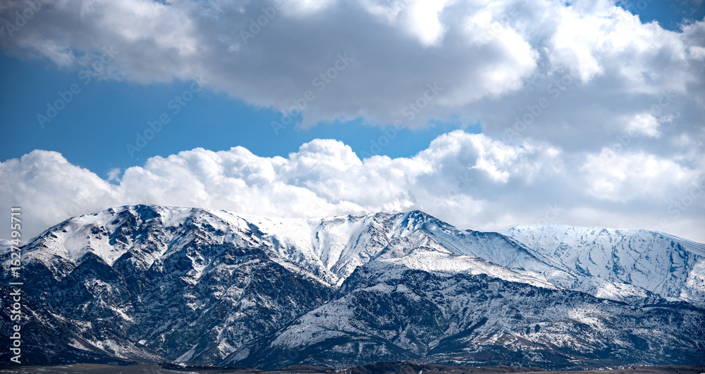 Fototapeta premium The image shows a majestic mountain range covered in snow under a dramatic sky filled with large, fluffy clouds. The mountains have a rugged and steep appearance, with various peaks and ridges visible