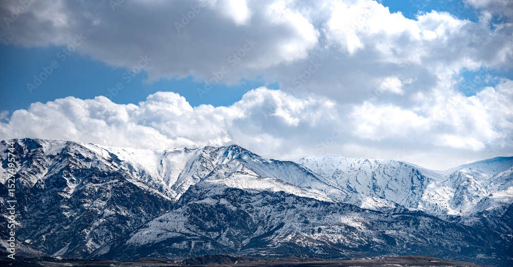 Fototapeta premium The image shows a majestic mountain range covered in snow under a dramatic sky filled with large, fluffy clouds. The mountains have a rugged and steep appearance, with various peaks and ridges visible