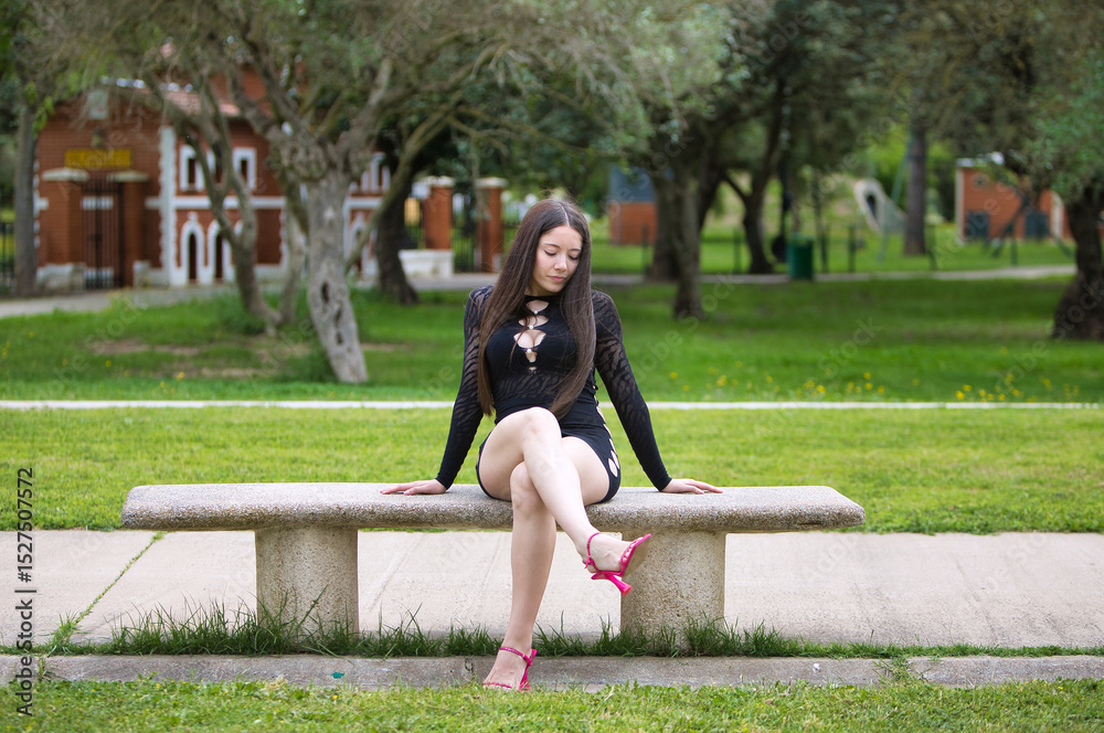 Fototapeta premium Latin woman, young and beautiful brunette dressed in black and pink heels sitting cross-legged on a park bench. The woman makes different expressions and gestures. Olive trees in the background.