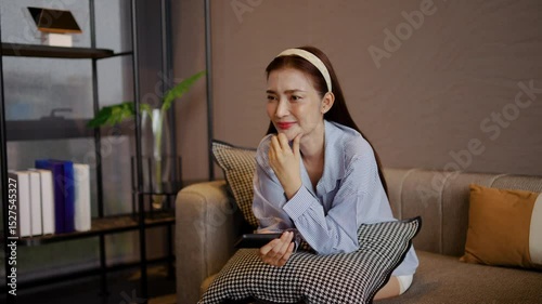 Relaxed adult asian woman sits comfortably on her living room sofa, engrossed in her favorite TV show or movie with remote in hand, as the evening unfolds peacefully.