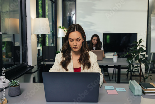 Young businesswoman working on laptop in modern office with colleagues in background