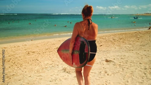 Old woman carries surfboard over head in hands. Senior female in swimsuit goes surfing on waves. Elderly lady beginner student goes on surfing session at tropical location at sunrise. Above head