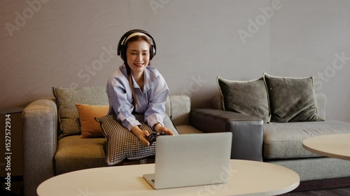 An adult asian woman enjoys her leisure time immersed in gaming by holding the joystick control via laptop on a comfortable sofa at cozy living room atmosphere. Her headphones enhance the fun