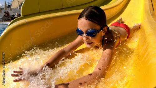 Schilderij op canvas Young girl of Latin ethnicity slides down a yellow water slide at an aqua park, wearing blue goggles and a red swimsuit