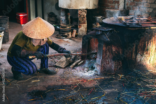 Obraz na plátně Vietnamese female stoker worker works in straw hat in workshop and lights a coal furnace at a reed dyeing plant in Vietnam