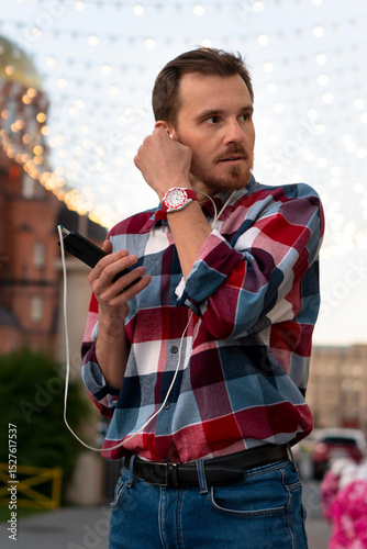 Man adjusting earphones while listening to music under lights. Guy with beard and modern hairstyle wearing in blue, red and white plaid shirt walking through the city streets in urban background