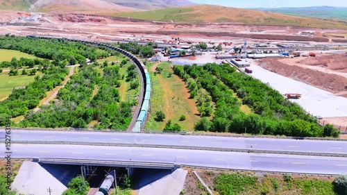 A freight train crosses a green valley near an industrial site. Aerial view of a highway, railway, and construction in the mountains.