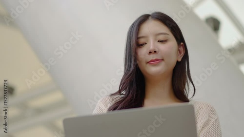 Portrait of attractive asian young woman sitting on a stairs on a city street working on her laptop. Typing on keyboard, Modern building with tall windows, Concept of freelance, browsing website