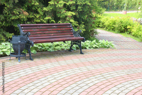 Fototapeta Naklejka Na Ścianę i Meble -  Wooden bench, wrought iron trash can near tile path in park. Green lawn, bushes and trees in summer on sunny day. Background for design. Ecology and nature. Landscape design. 