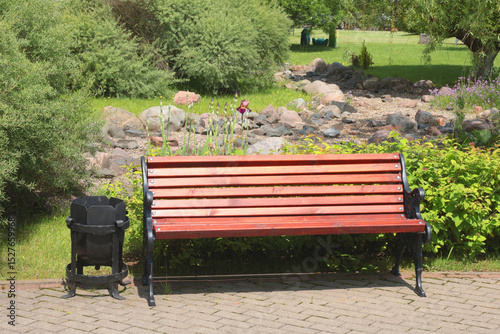 Fototapeta Naklejka Na Ścianę i Meble -  Wooden bench, wrought iron trash can near tile path in park. Green lawn, bushes and trees in summer on sunny day. Background for design. Ecology and nature. Landscape design. 