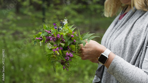 Woman holding a wildflower bouquet in a green forest, wearing a smartwatch and grey clothes, with focus on hands and flowers.
