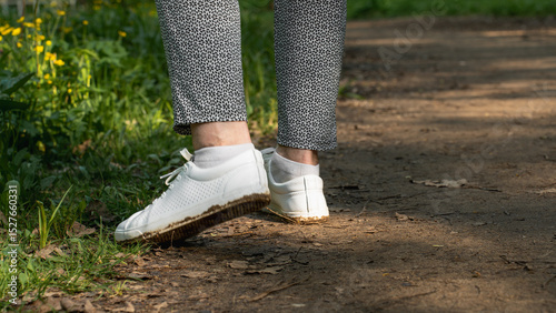 Close-up of a person's legs walking on a forest path wearing white sneakers and patterned pants, surrounded by green grass and yellow wildflowers in spring sunlight.