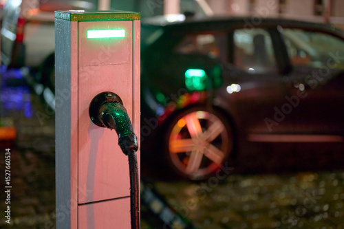 electric vehicle charging station at night with car plugged in, showcasing modern eco-friendly technology for sustainable urban transportation. green lights illuminate wet pavement and charger. photo