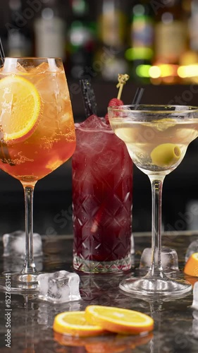 A static, eye-level medium close-up video of three vibrant cocktails: orange spritz, berry, and classic martini, garnished, on a wet, reflective bar counter with soft, warm bokeh of liquor bottles.