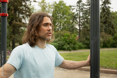 Portrait of tired sporty caucasian man thoughtful expression, lost in thought at public park near sport ground equipment. Laziness, forces himself to exercise. Slow living. Outdoor