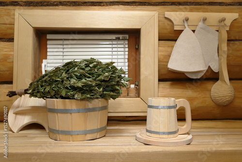 A wooden beer mug stands on tray next to bucket with dry birch broom on plank bench in the sauna interior in front of the window with blinds, which is in log wall with set of sauna accessories on it.