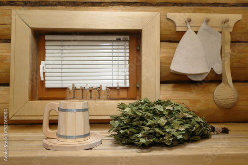 A wooden beer mug stands on a tray next to a dry birch broom on a plank bench in the sauna interior in front of the window with blinds, which is in a log wall with a set of sauna accessories on it.