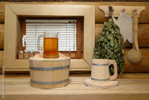 A glass mug of light beer stands on a wooden bucket on a plank bench in the sauna interior in front of the window with blinds, which is in a log wall with a set of sauna accessories on it.