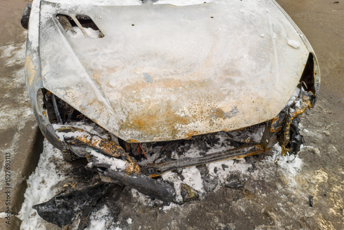 Car is covered in snow and has a black hood. The hood is missing and the car is burnt
