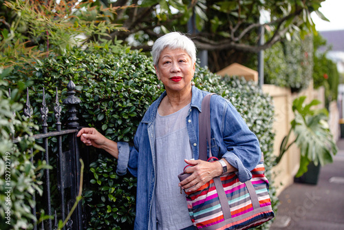 Senior Asian woman standing next to a black wrought iron fence and a lush green hedge