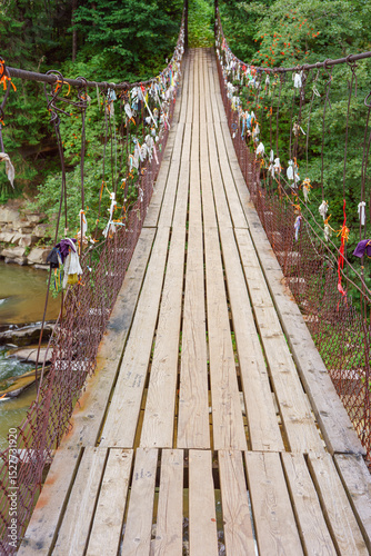 Wooden suspension bridge with colorful ribbons hanging from wires crosses fast mountain river, mountain creek in lush green coniferous Carpathian forest. Adventure, travel, hiking and local charm.