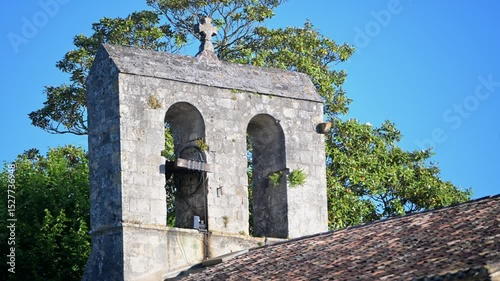 A charming bell tower rises against a clear blue sky, surrounded by vibrant greenery. The structure features two bells, inviting exploration of its rich history and picturesque surroundings.