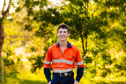 Portrait of happy young tradesman apprentice in high vis workwear against leafy green bokeh backdrop