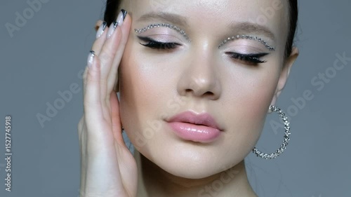 Female with unusual rhinestones makeup and earrings in the ears. Hand near face.
