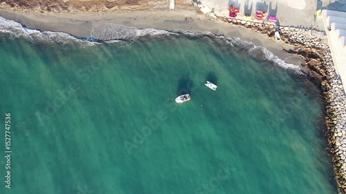 Upward-moving drone shot above Durres Beach, Albania, revealing boats anchored offshore, gentle waves on the shoreline, colorful umbrellas, and a geometric coastal promenade from a top-down view.